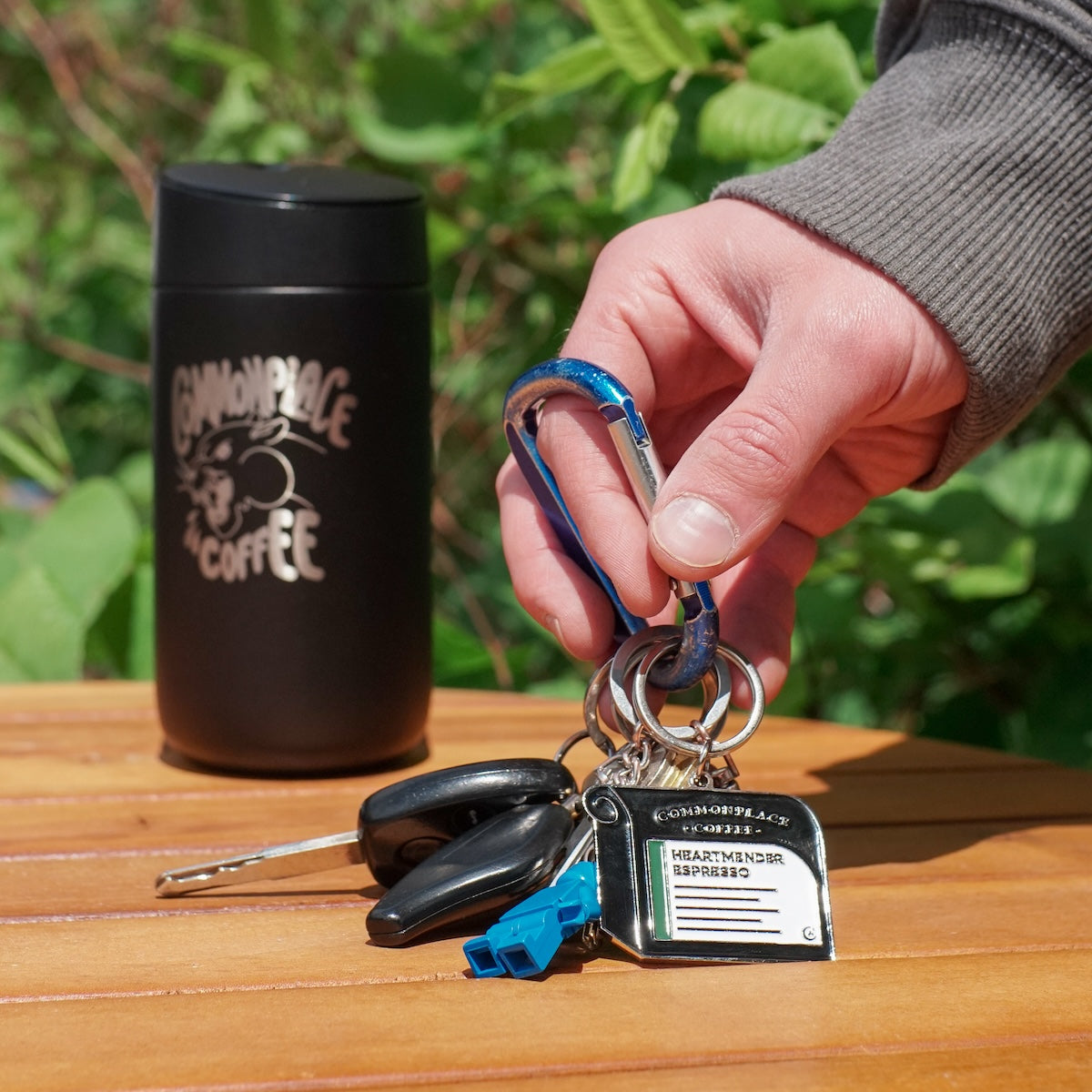 A hand picking up a carabiner off of a wooden table outside. On the carabiner is a keychain designed to look like a bag of Commonplace Coffee, car keys, house keys, and a small Lego keychain. Further bag on the table is a travel mug that says 
