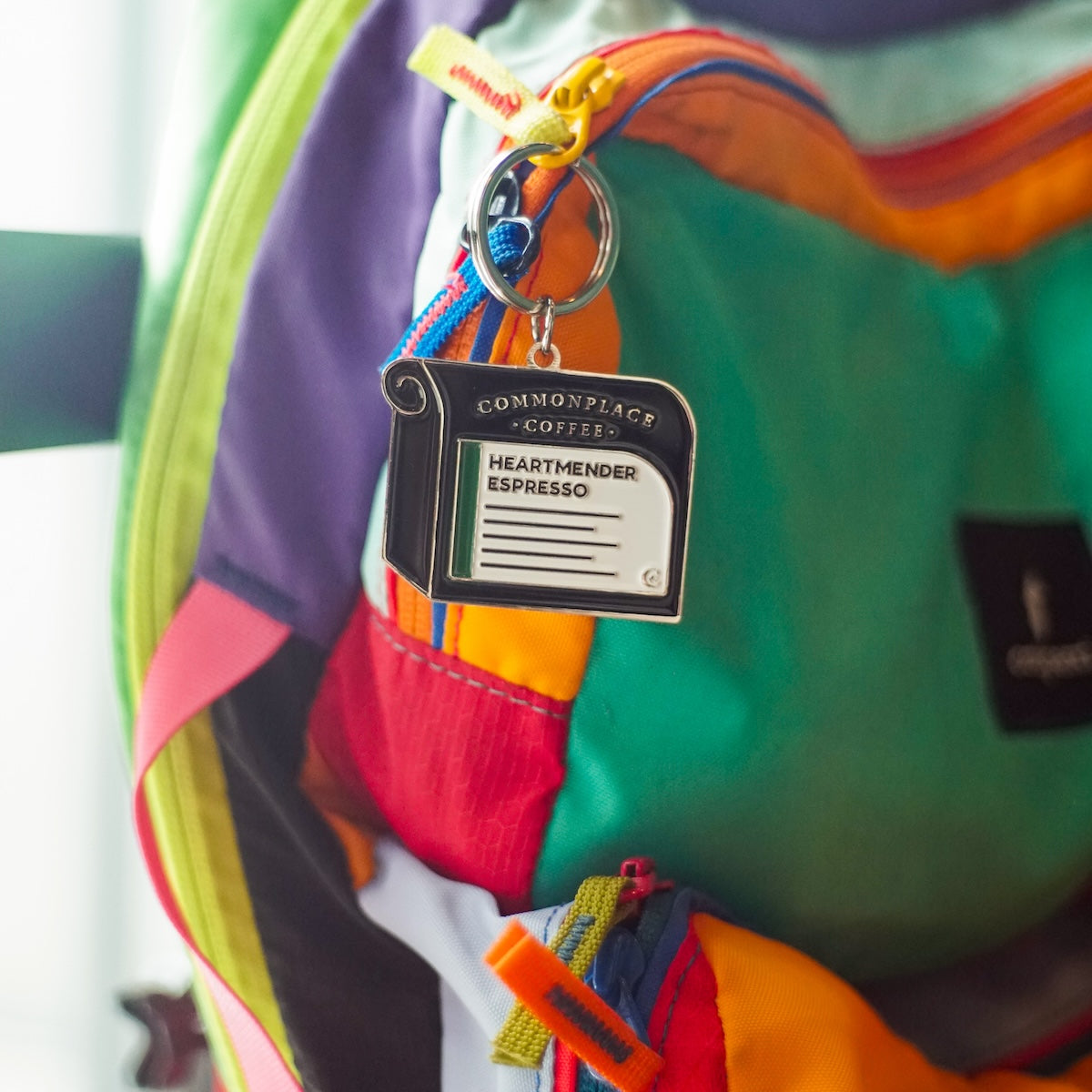 A keychain designed to look like a bag of Commonplace Coffee's Heartmender Espresso hangs on the zipper of a colorful backpack.